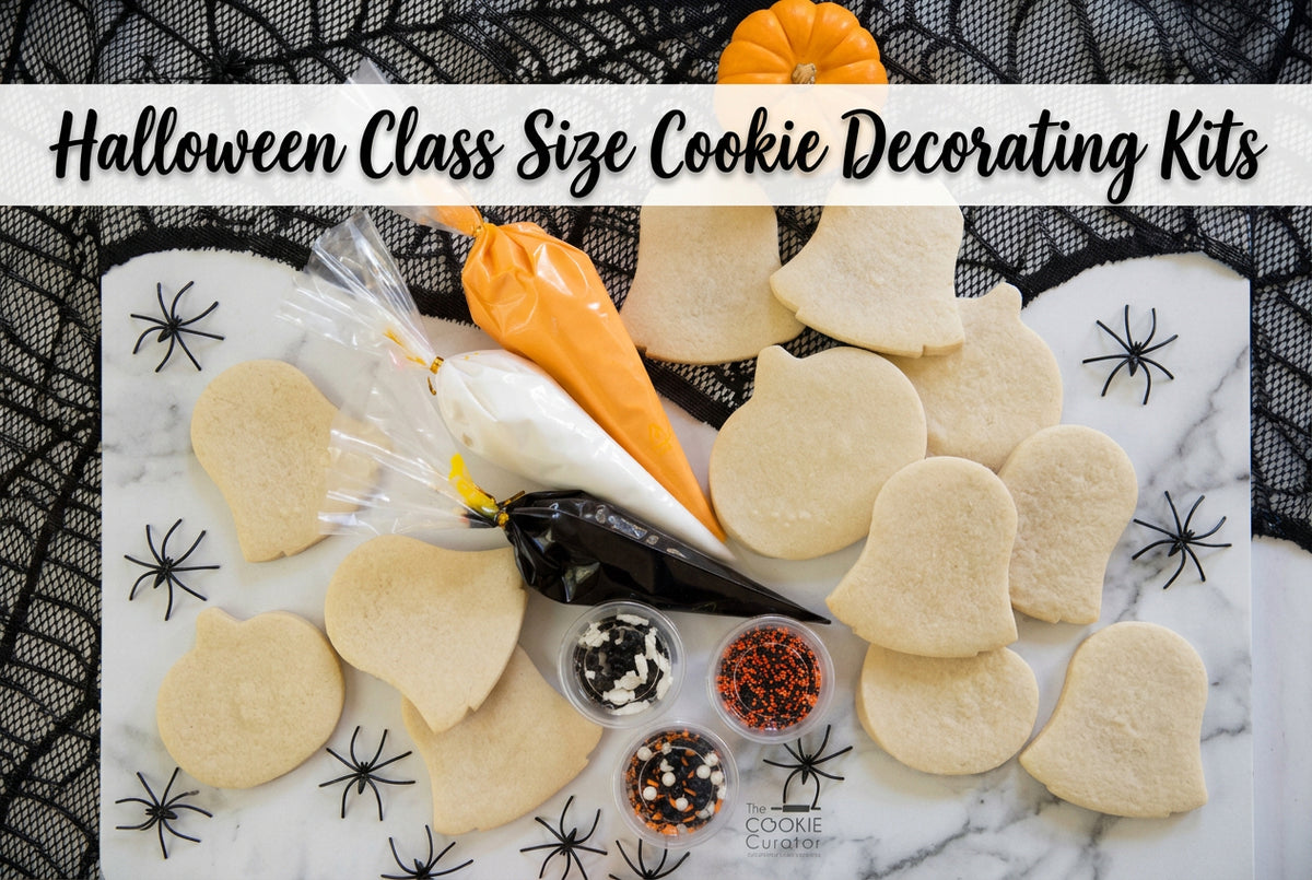 Sugar cookies shaped like ghosts and bats with bags of royal icing and containers of sprinkles. The cookies are displayed on a table with Halloween decorations including black spider webs and a small orange pumpkin.